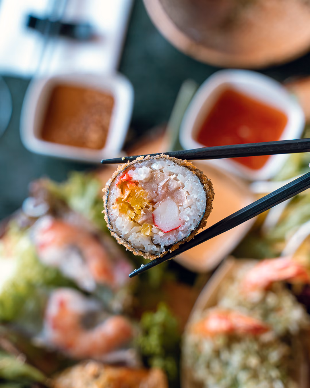 Ein frittiertes Maki-Röllchen mit Surimi und Gemüse zwischen schwarzen Essstäbchen, im Hintergrund unscharf zwei Dip-Schälchen.