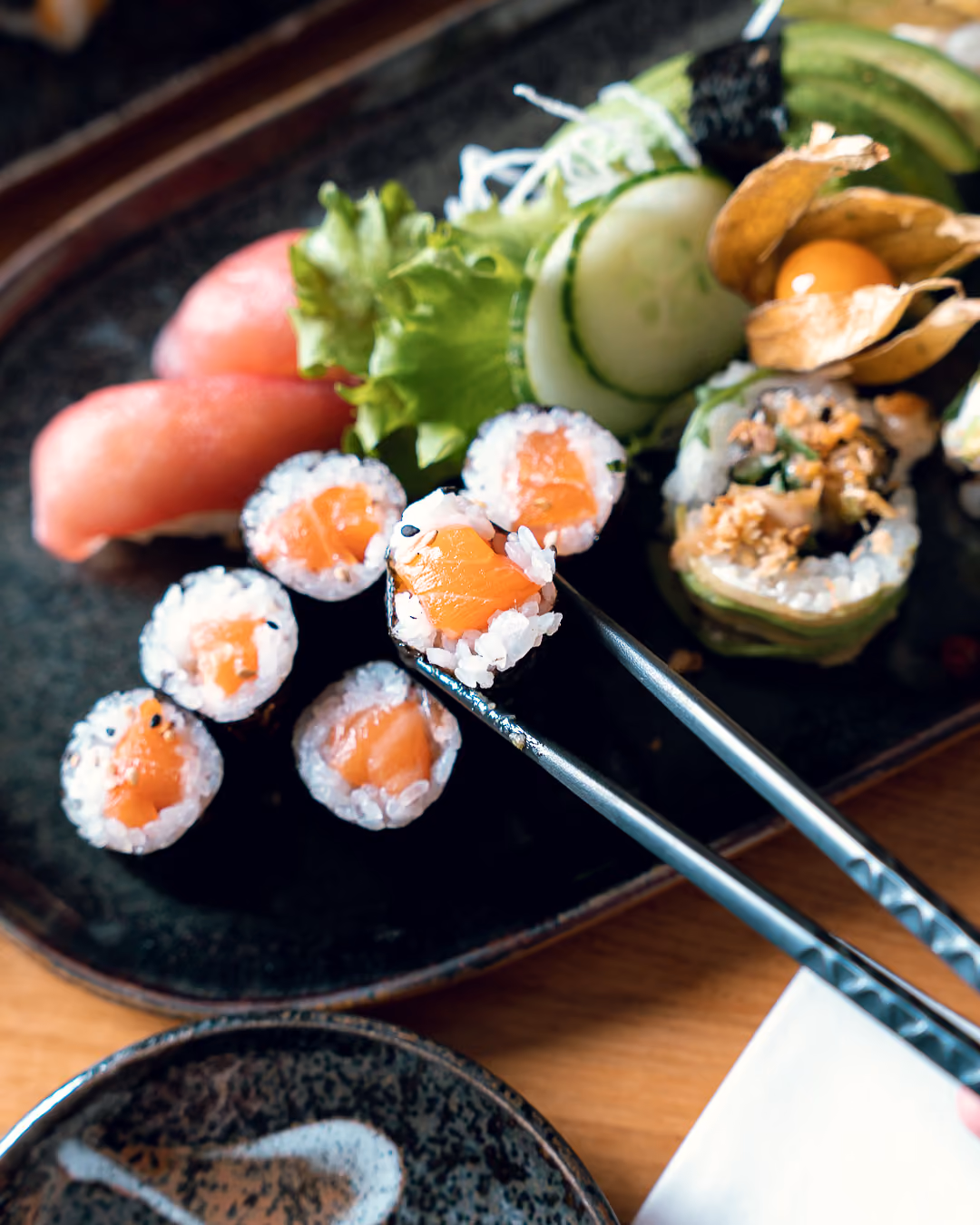 Ein Lachs-Maki wird mit Essstäbchen von einer dunklen Keramikplatte gehoben, dahinter Thunfisch-Nigiri und Avocado-Rollen.