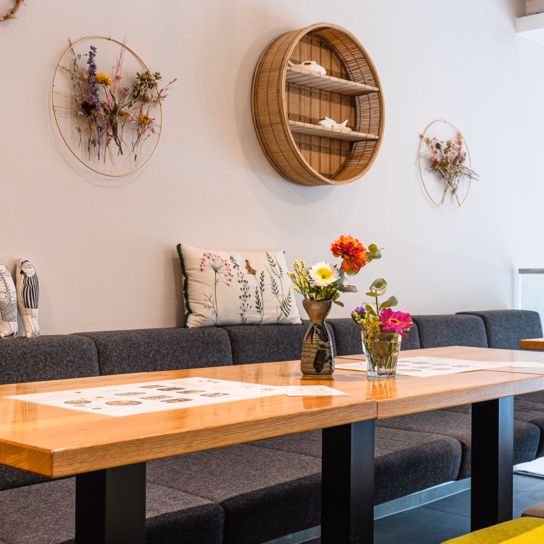 Lange Holztafel mit Speisekarten und Blumenvasen vor der grauen Sitzbank im Gastraum der Bento Boutique.