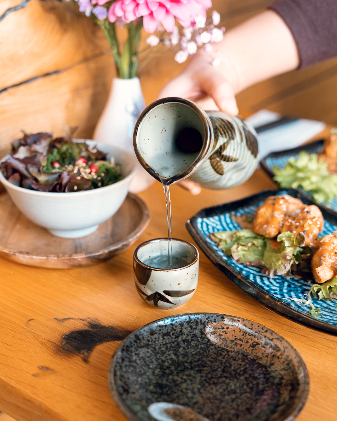 Eine Hand gießt Sake aus einer Keramikkanne in ein kleines Schälchen, daneben Salat und ein Teller mit frittiertem Hähnchen.
