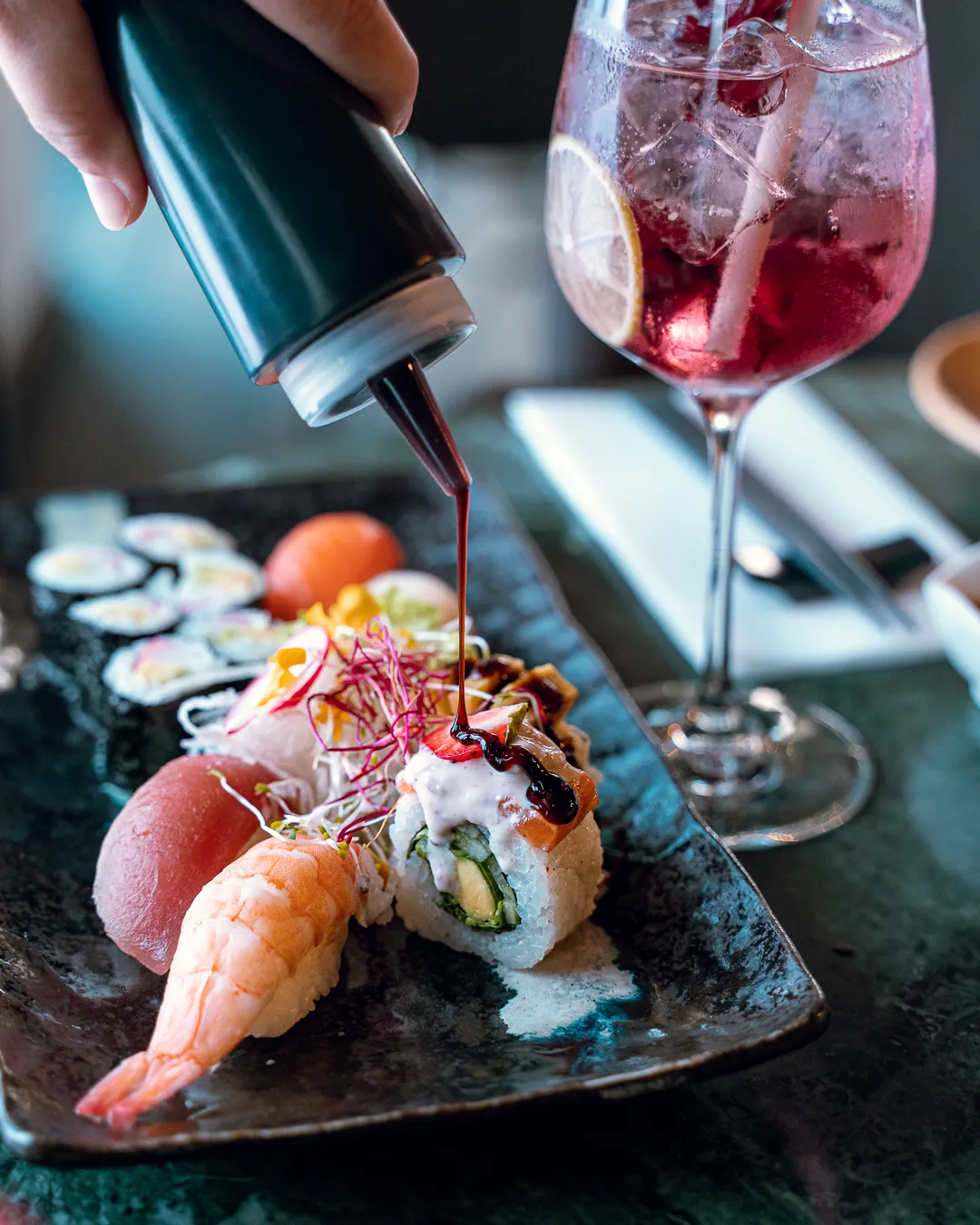 Dunkle Schieferplatte mit Nigiri, Maki und einer Inside-out-Rolle; eine Hand träufelt aus einer kleinen Flasche dunkle Sauce darüber, daneben steht ein Glas mit fruchtigem Getränk