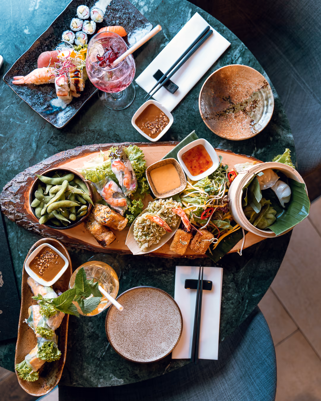 Aufsicht auf einen Marmortisch mit Vorspeisenbrett aus Edamame, Sommerrollen und Dumplings, dazu Sushi-Teller und zwei Cocktails.